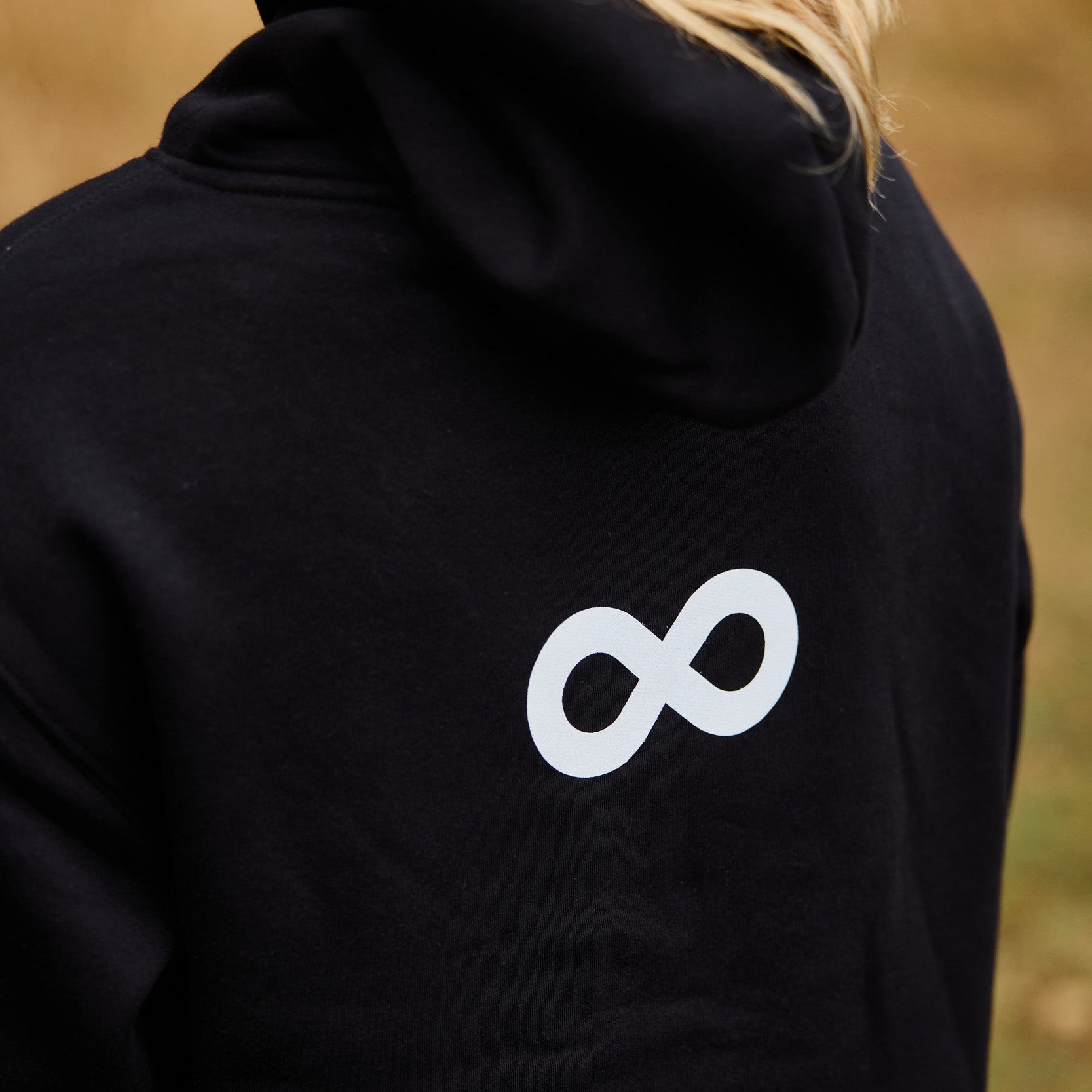 Person wearing a black hoodie with a white infinity symbol on the back, standing in a natural setting.