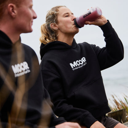 Two people wearing black hoodies with 'MOOR' branding, one drinking from a pink bottle outdoors.