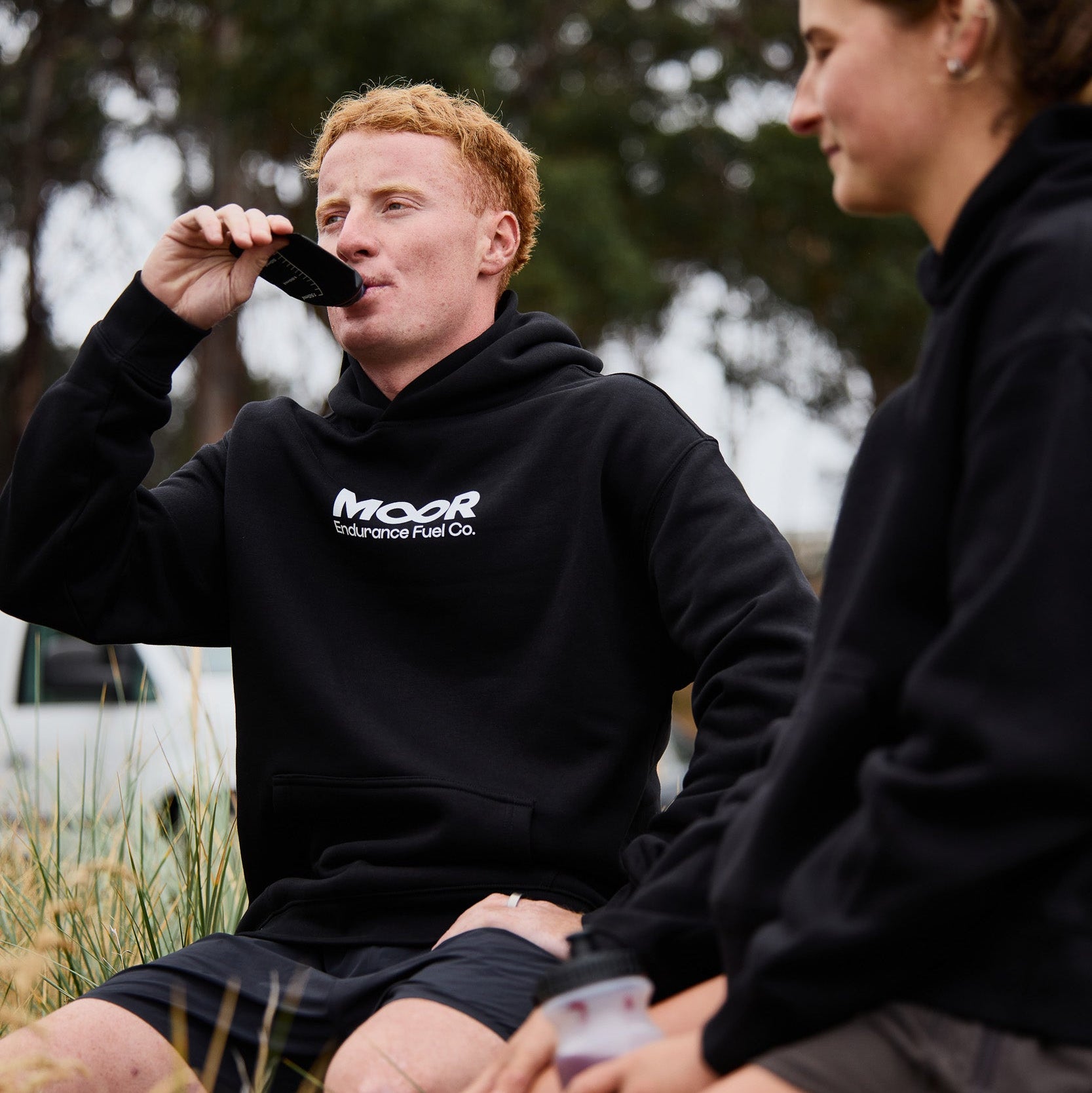 Two people sitting outdoors, one wearing a black hoodie with 'MOOR' branding.